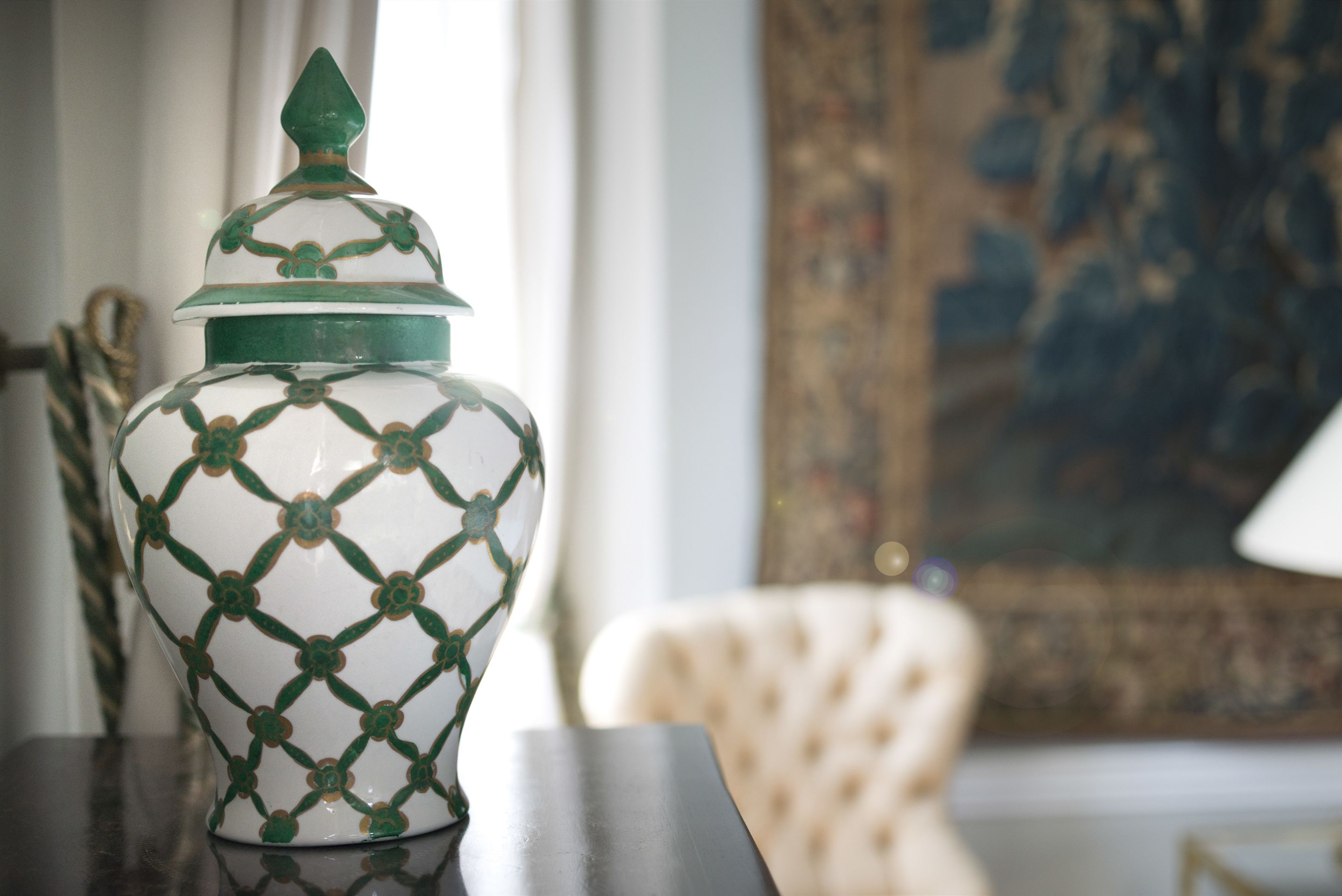A decorative ceramic jar with a green and white pattern, placed on a table in a room with a white chair and a painting in the background.