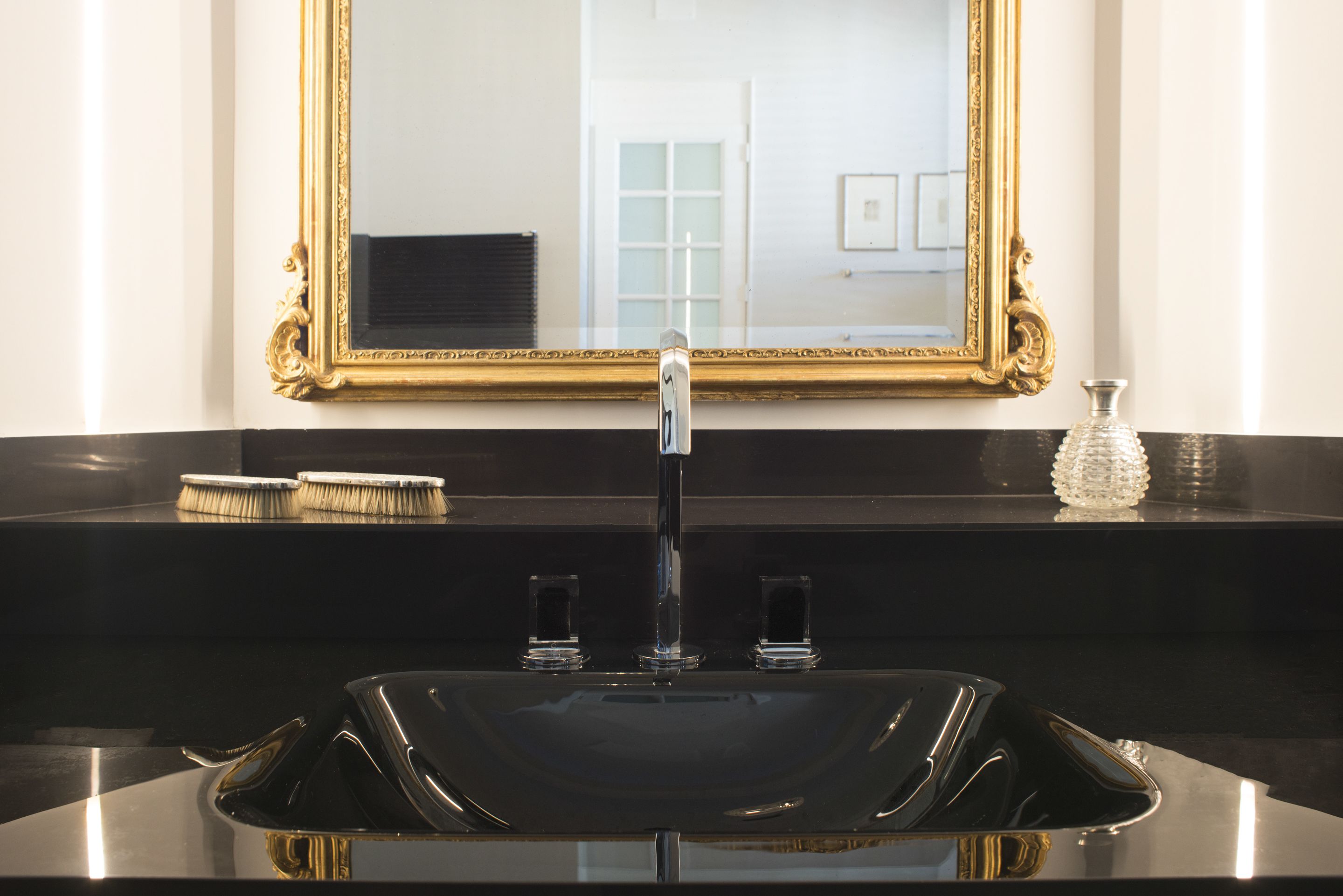 Une salle de bain avec un lavabo noir, un miroir à cadre doré et un vase en verre posé sur le comptoir.