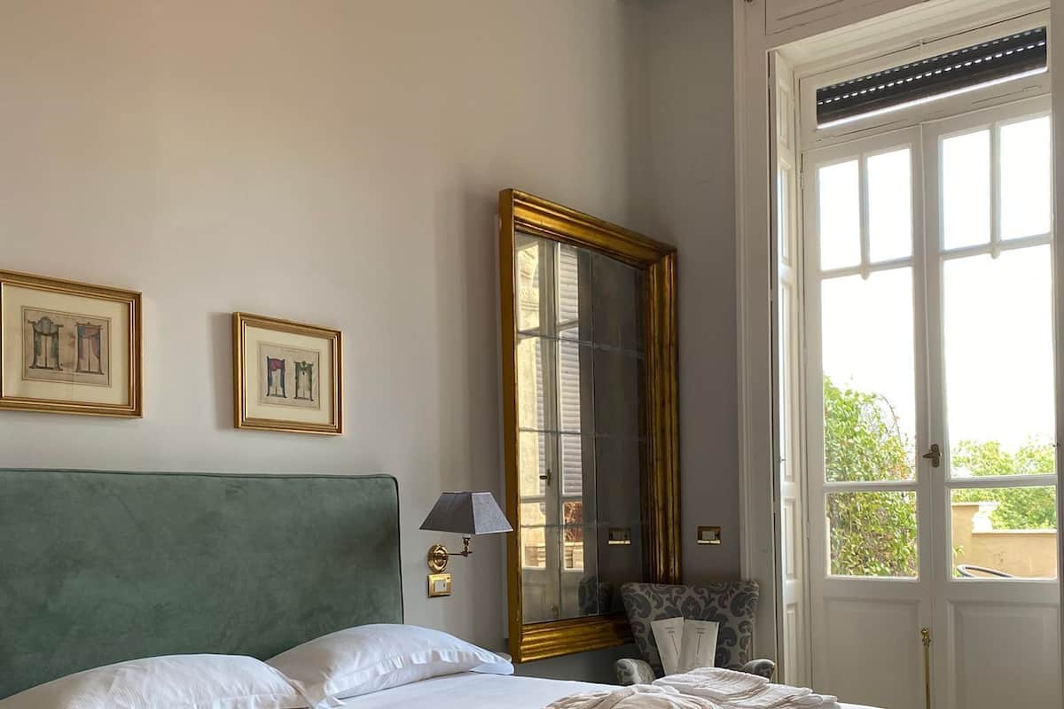 Un lit bien fait, avec du linge de lit blanc, une tête de lit recouverte d’un tissu vert et un grand miroir au mur.