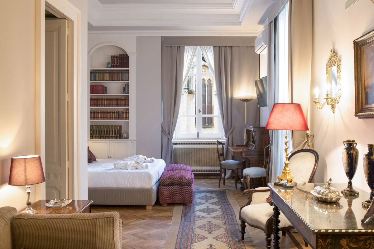 Une chambre d’hôtel avec un lit, un bureau avec une lampe, une bibliothèque et une fenêtre avec des rideaux.