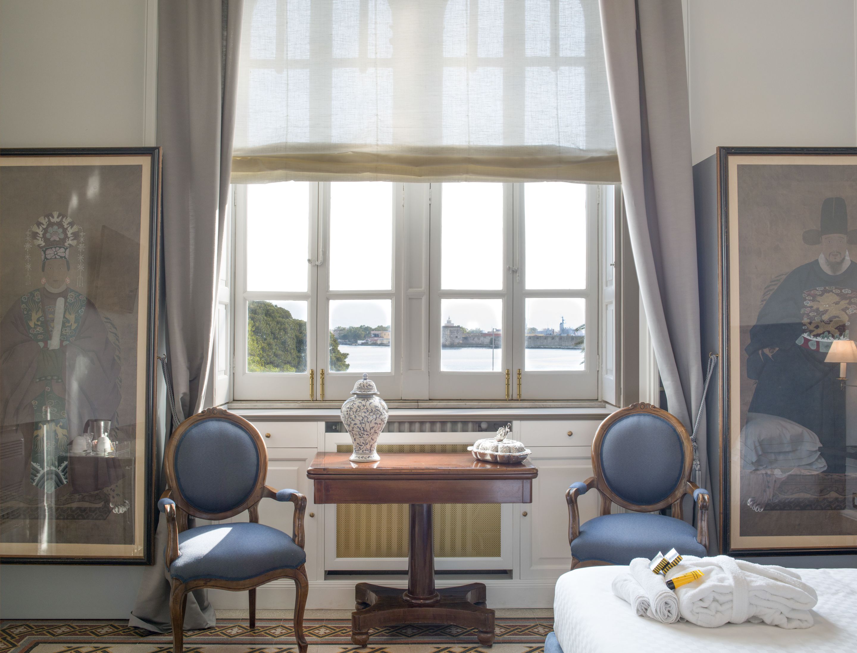 Ein Hotelzimmer mit großem Fenster, zwei Stühlen, einem kleinen Tisch mit einer Vase und Bildern an den Wänden.