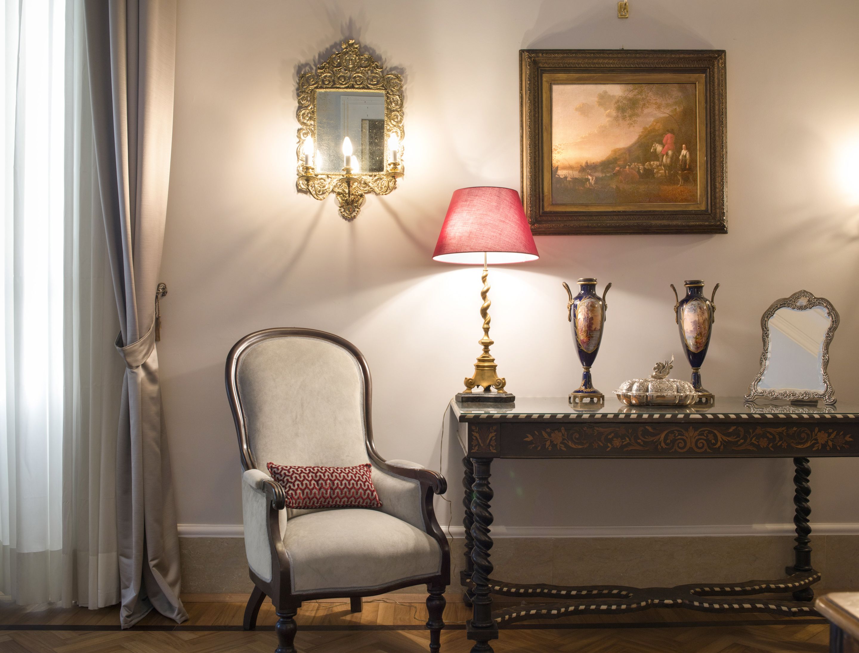 Una habitación con una silla, una mesa con objetos decorativos, un espejo adosado a la pared y un cuadro enmarcado.