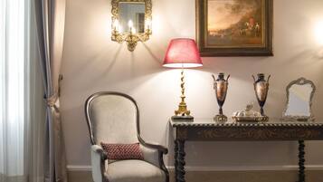 Una habitación con una silla, una mesa con objetos decorativos, un espejo adosado a la pared y un cuadro enmarcado.