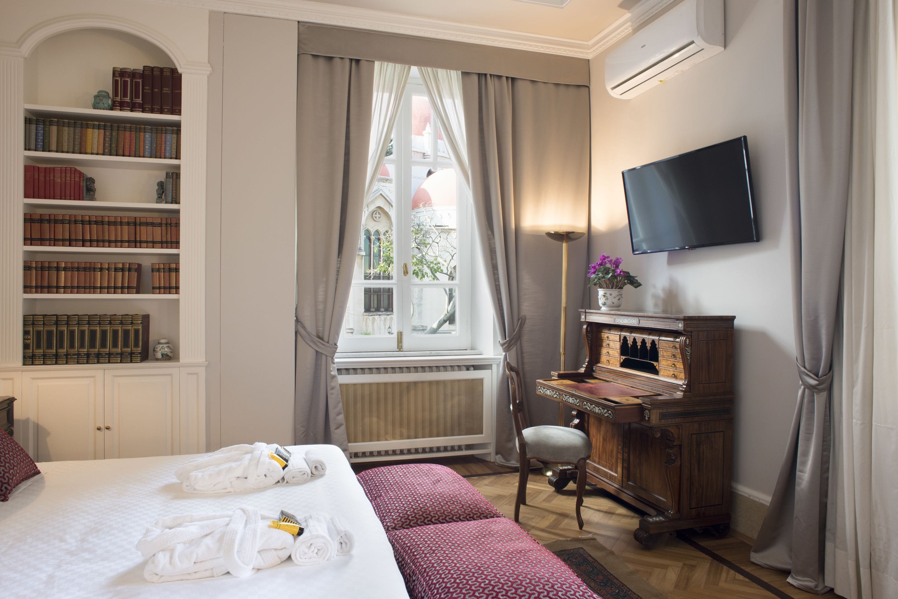 A hotel room with a large bed, a bookshelf, a desk with a piano, a television, and a window with curtains.