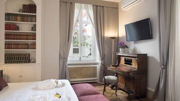 A hotel room with a large bed, a bookshelf, a desk with a piano, a television, and a window with curtains.