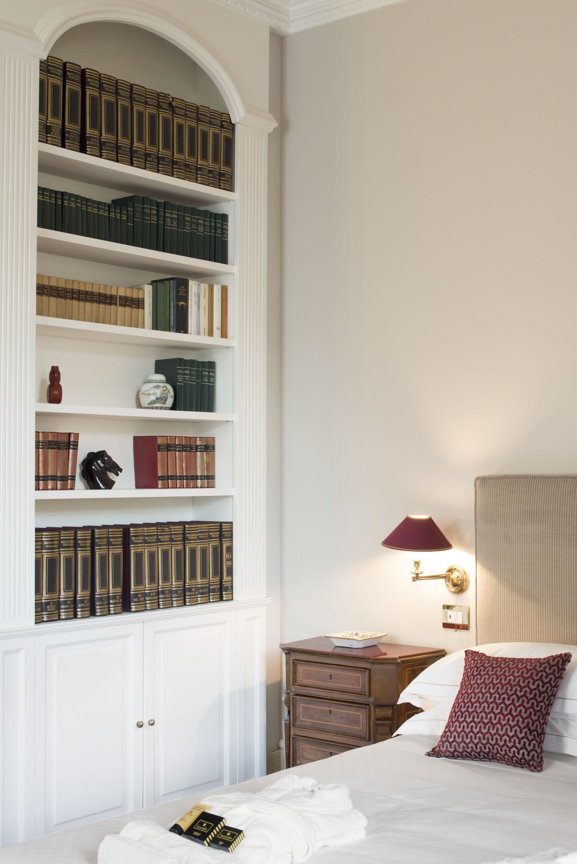 Un dormitorio con una amplia estantería llena de libros, una mesita de noche con lámpara y una cama con un cojín estampado.