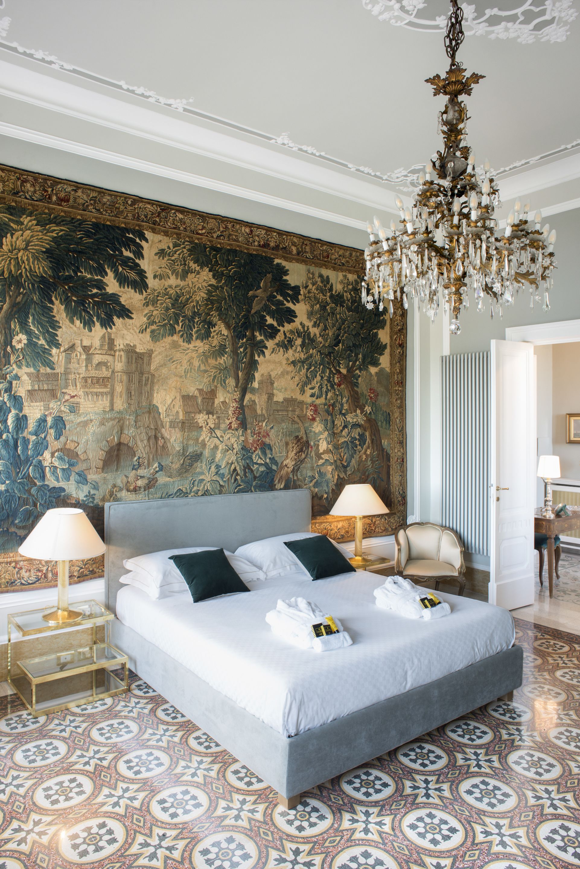 A spacious bedroom with a large bed, ornate chandelier, and a detailed tapestry on the wall.