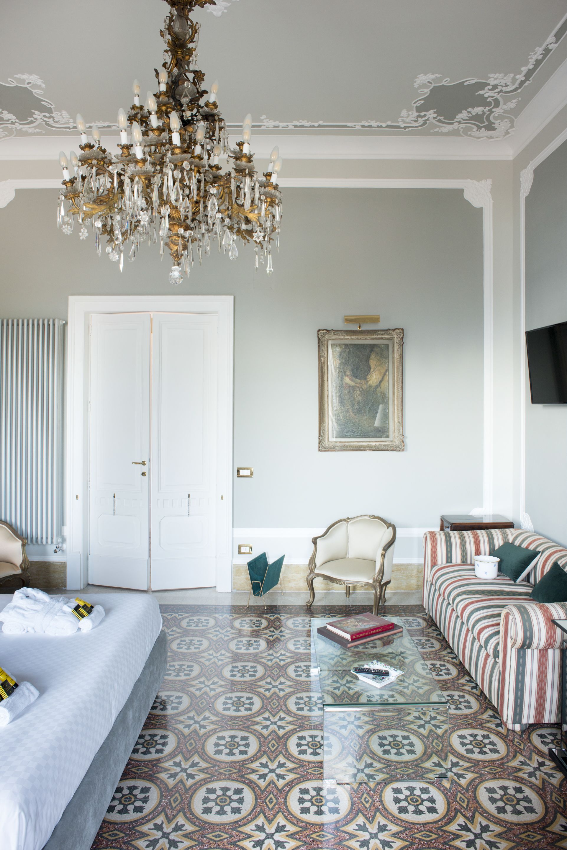 Une chambre d’hôtel avec un grand lit, un lustre, un sol à motifs, un canapé, un fauteuil et un tableau au mur.