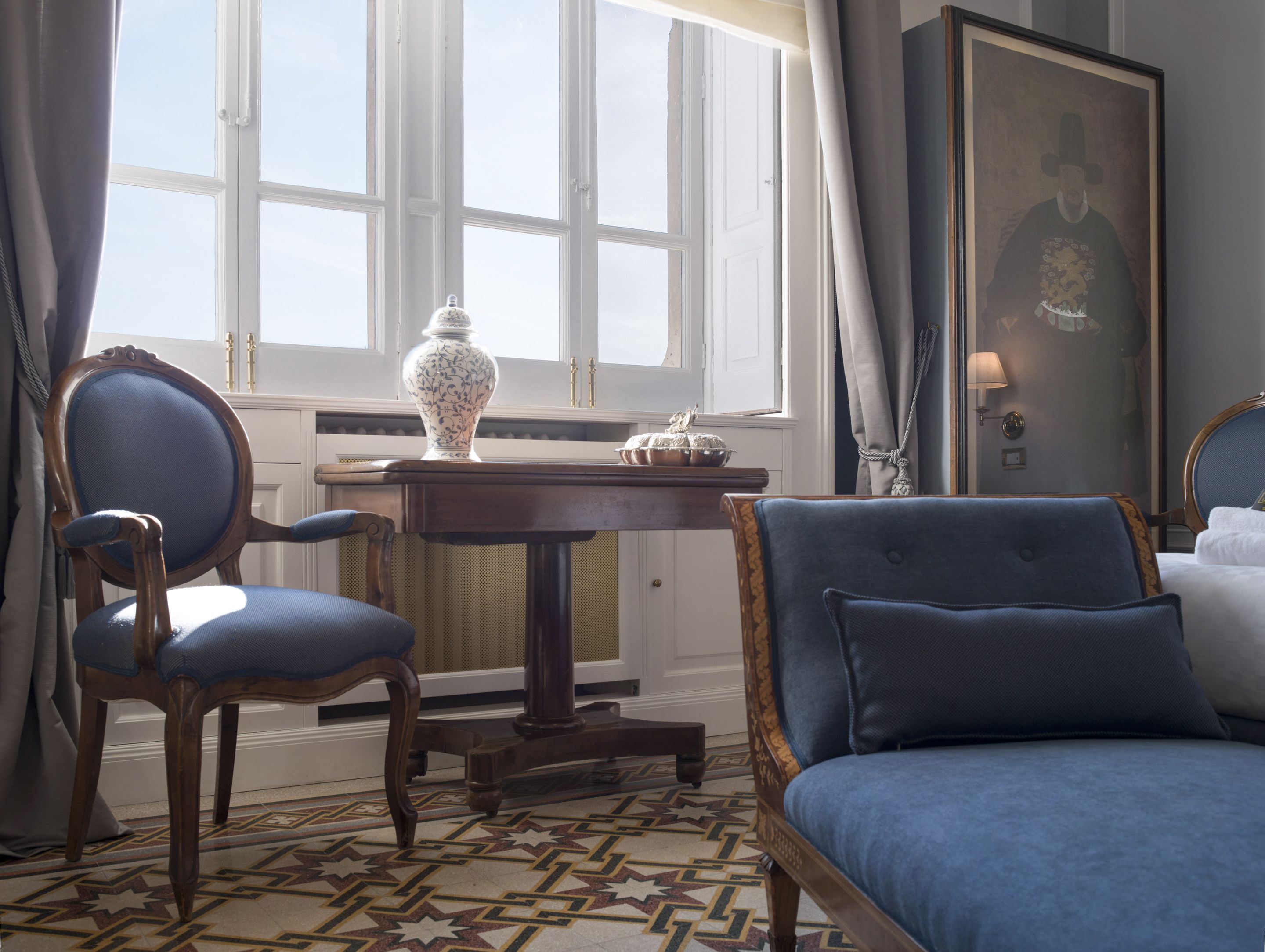 A hotel room with a large window, a table with a vase, two chairs, a bed, and a painting on the wall.