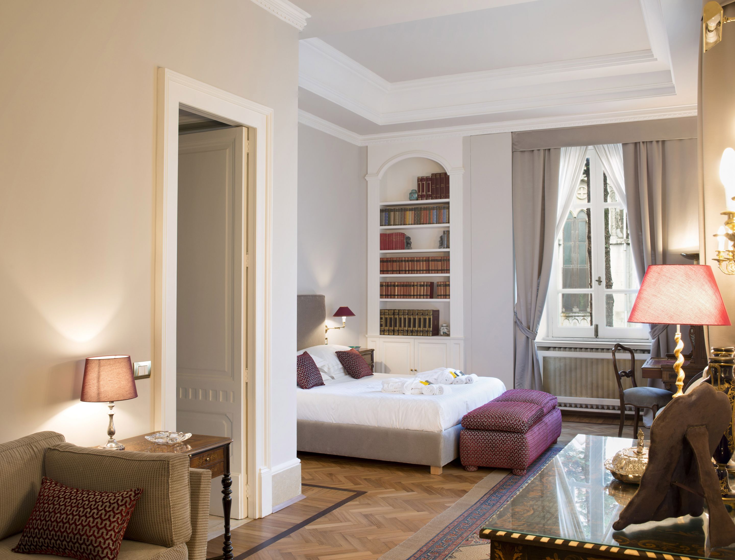 A hotel room with a bed, bedside table, lamp, bookshelf, and a window with curtains.