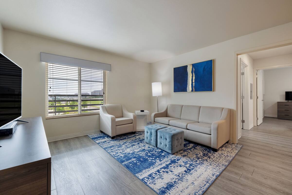 A modern living room with a sofa, armchairs, a coffee table, and a TV. There is a large window with blinds, a blue patterned rug, and a painting on the wall.