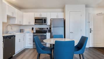 A modern kitchen with white cabinets, a stainless steel refrigerator, a microwave, and a dishwasher. There is a round table with four blue chairs.