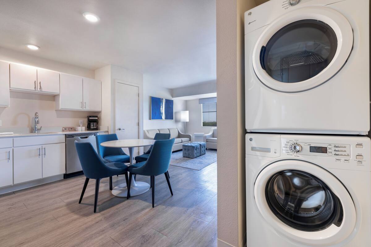 A modern kitchen with white cabinets, a dishwasher, and a coffee maker. There is a dining area with a round table and blue chairs. A washing machine is visible in the foreground.