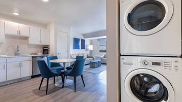 A modern kitchen with white cabinets, a dishwasher, and a coffee maker. There is a dining area with a round table and blue chairs. A washing machine is visible in the foreground.