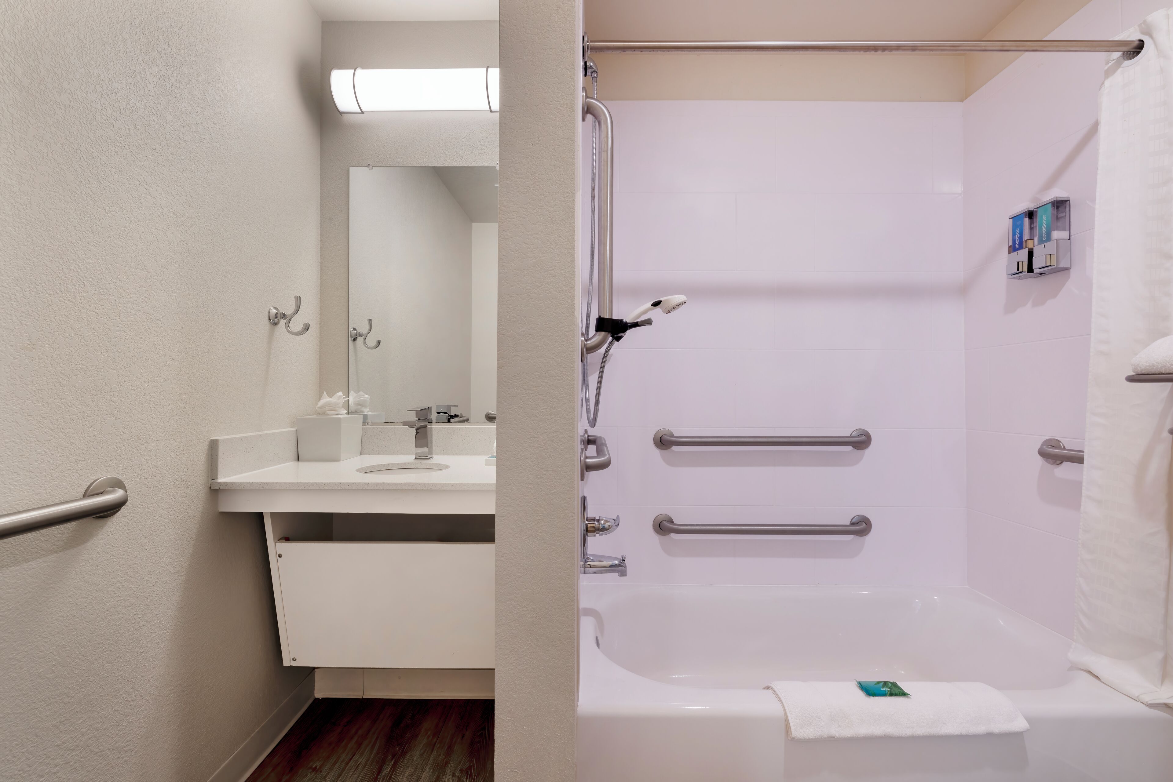 Une salle de bain moderne avec un lavabo, un miroir et une baignoire blancs. Elle est équipée de barres d’appui pour plus de sécurité et d’un porte-serviettes.