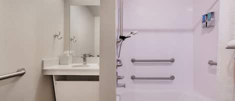 Une salle de bain moderne avec un lavabo, un miroir et une baignoire blancs. Elle est équipée de barres d’appui pour plus de sécurité et d’un porte-serviettes.