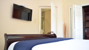 A hotel room with a bed, a television mounted on the wall, and an open door leading to another room. - The Barcelona by Pro Homes Jamaica (Kingston)