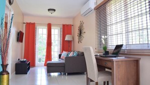 A modern living room with a sofa, a desk with a laptop, a lamp, and a large window with blinds. - The Barcelona by Pro Homes Jamaica (Kingston)