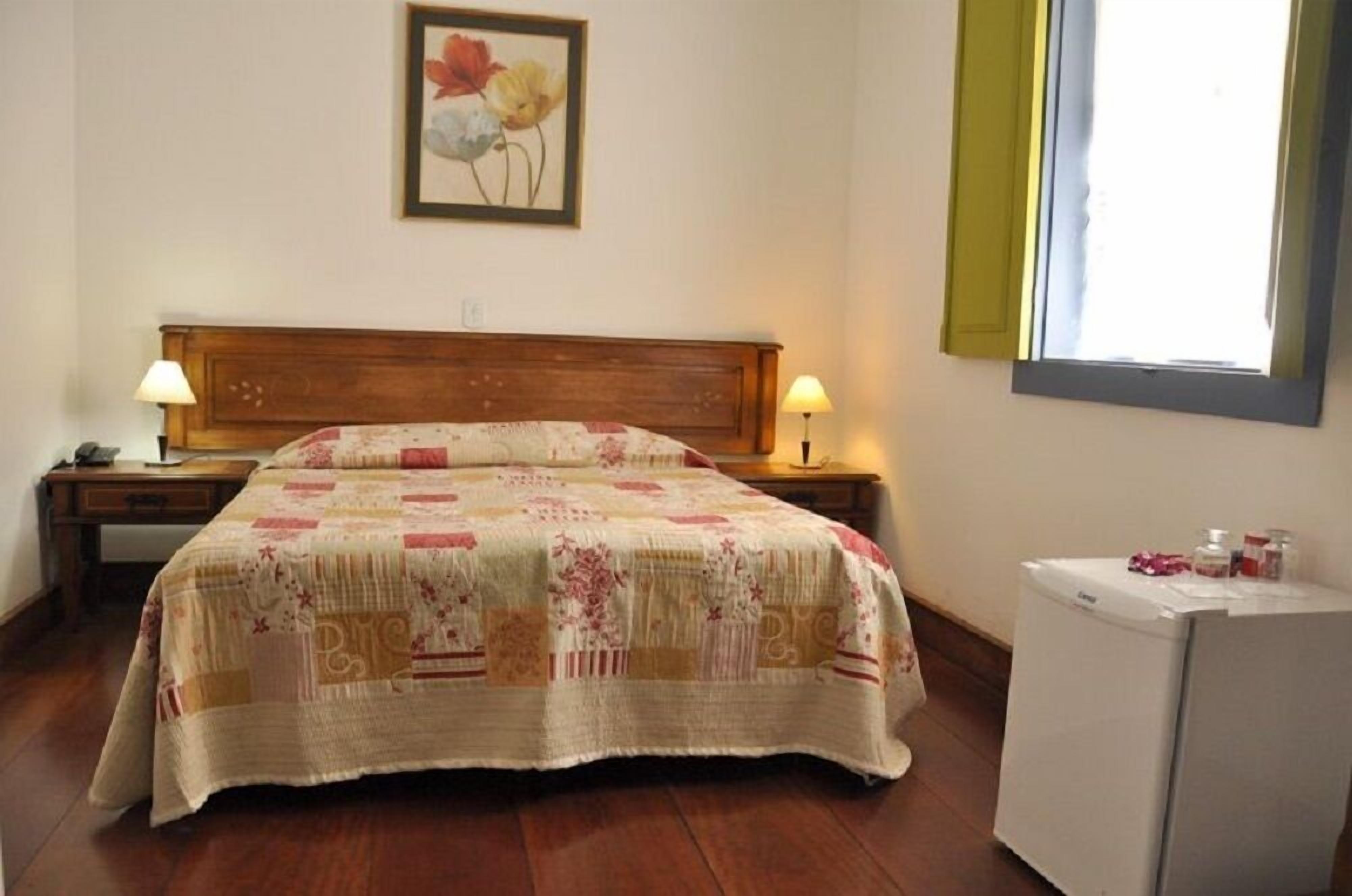 A bedroom with a wooden headboard, a floral bedspread, two bedside lamps, a nightstand, a small table with a lamp, a window with blinds, and a refrigerator.