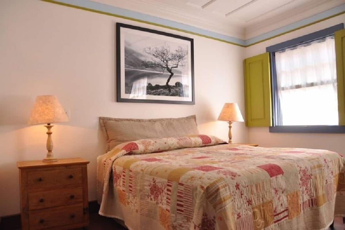 A bedroom with a bed, two bedside tables with lamps, a framed picture on the wall, and a window with curtains.