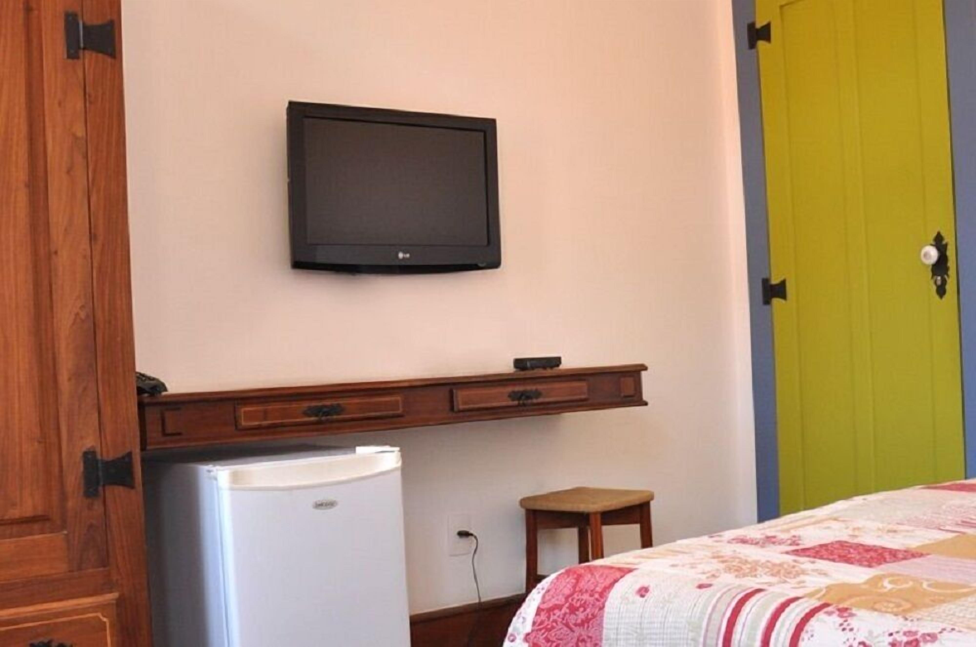 Una habitación con una televisión montada en la pared, un taburete pequeño de madera, un refrigerador blanco y una cama con una colcha estampada.