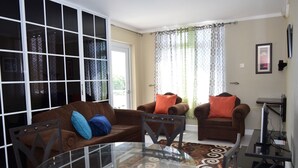 Una sala de estar con una mesa de vidrio, dos sofás marrones con cojines naranjas y un televisor sobre un mueble.