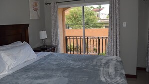 Un dormitorio con cama, una mesita de noche con lámpara, una ventana con cortinas y vistas a un balcón con barandilla.