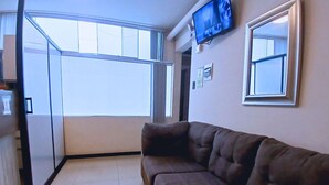 A living room with a brown sofa, a television mounted on the wall, and a large window providing natural light.