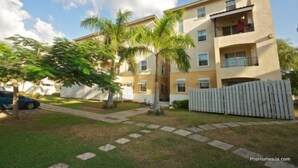 A multi-story residential building with a balcony on the second floor, a fenced yard, and a palm tree. - Winchester 12A by Pro Homes Jamaica (Kingston)