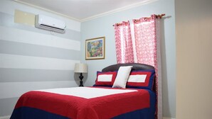 A hotel room with a bed, a nightstand, a lamp, an air conditioning unit, and a painting on the wall. - Winchester 12A by Pro Homes Jamaica (Kingston)