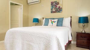 A bedroom with a bed, two bedside lamps, a wall-mounted air conditioner, and a colorful artwork. - Winchester 07A by Pro Homes Jamaica (Kingston)