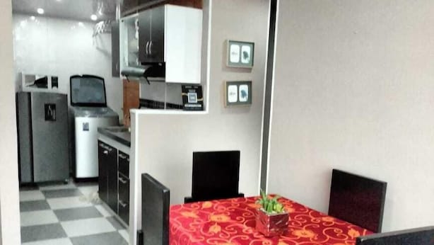 Un comedor compacto con mantel rojo estampado, sillas negras y una pequeña planta en maceta. Se observa una kitchenette con microondas y refrigerador al fondo.