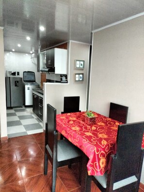 Un comedor compacto con mantel rojo estampado, sillas negras y una pequeña planta en maceta. Se observa una kitchenette con microondas y refrigerador al fondo.