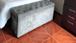 Un dormitorio con cama, un banco tapizado en gris, un armario y suelo de baldosas.