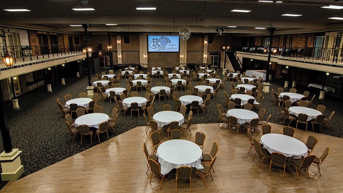 A large banquet hall with multiple round tables and chairs, a stage with a screen, and a balcony overlooking the room.