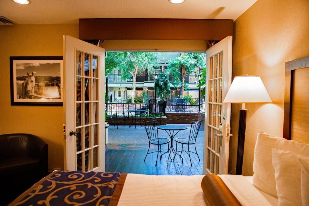 Habitación de hotel con puerta abierta a un patio exterior con mesas y sillas, una cama con almohada y una lámpara.