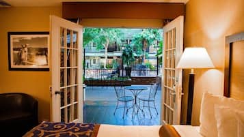Habitación de hotel con puerta abierta a un patio exterior con mesas y sillas, una cama con almohada y una lámpara.