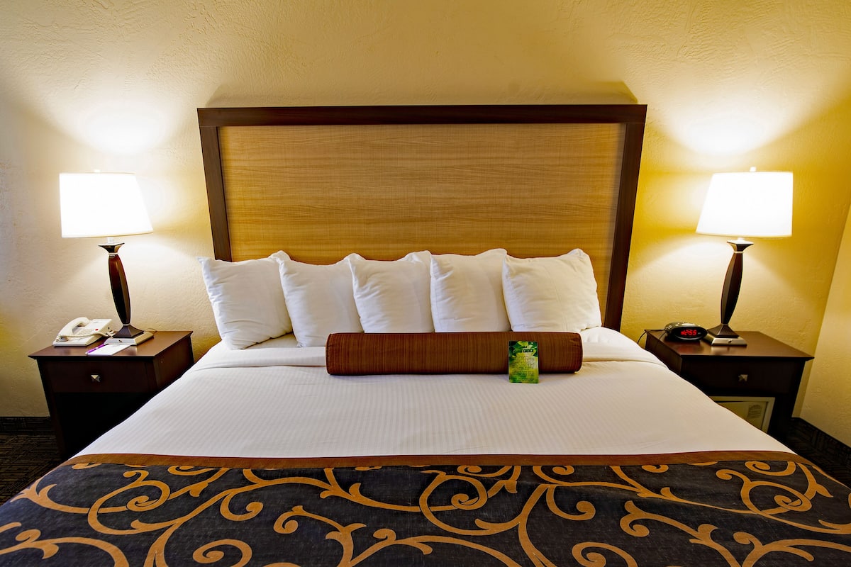 A hotel room with a large bed, two bedside tables, a nightstand, a lamp, a clock, and a green bottle on the bed.