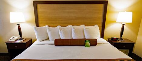 A hotel room with a large bed, two bedside tables, a nightstand, a lamp, a clock, and a green bottle on the bed.