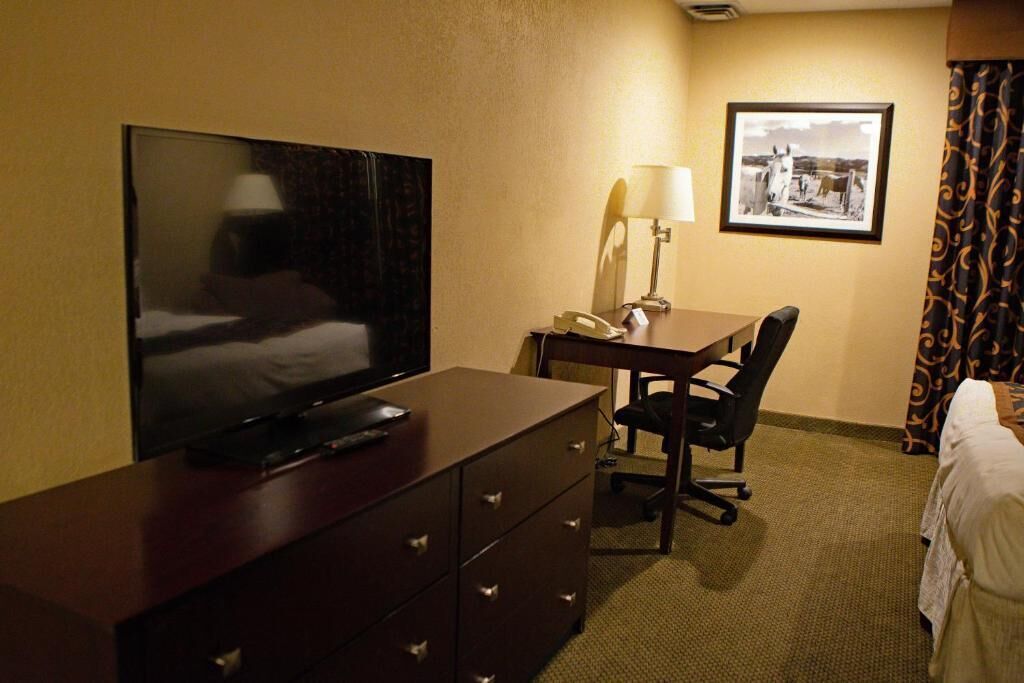 Habitación de hotel con televisión de pantalla plana sobre un tocador, un escritorio con lámpara, una silla y una cama con cortinas.