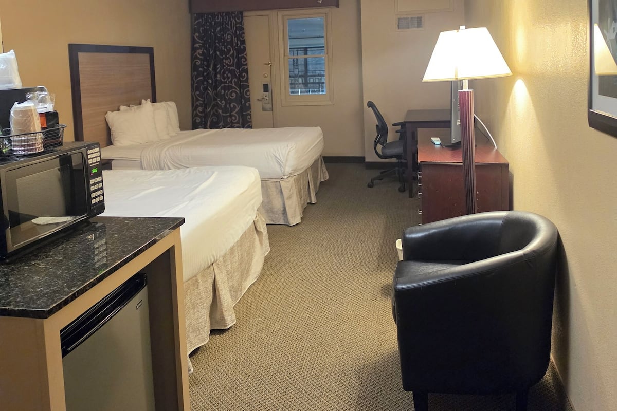 A hotel room with two beds, a desk with a microwave, a chair, a desk lamp, and a window with curtains.
