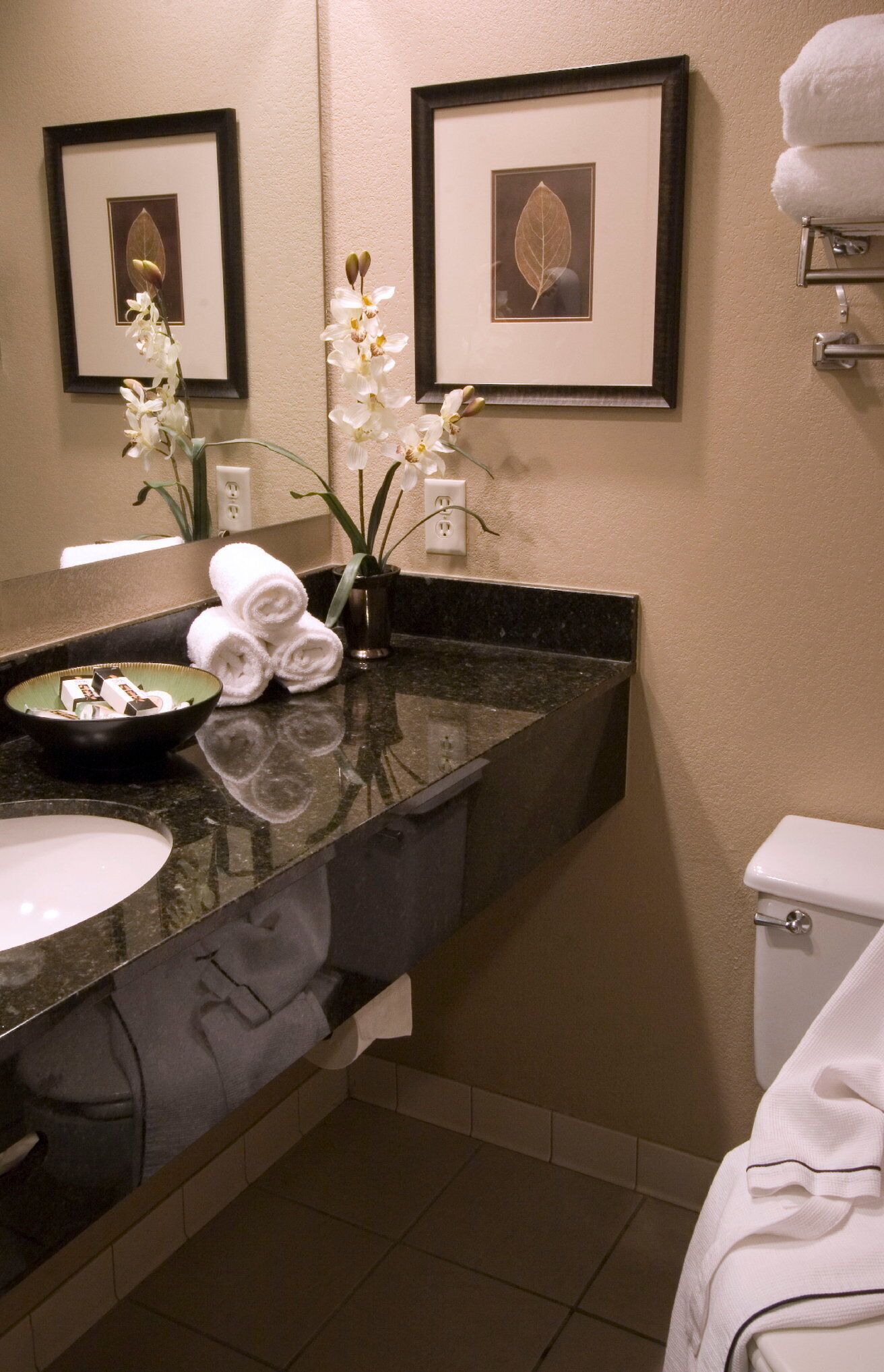 A bathroom with a black marble countertop, white sink, and a toilet. There are framed pictures on the wall and a vase with white flowers on the counter.