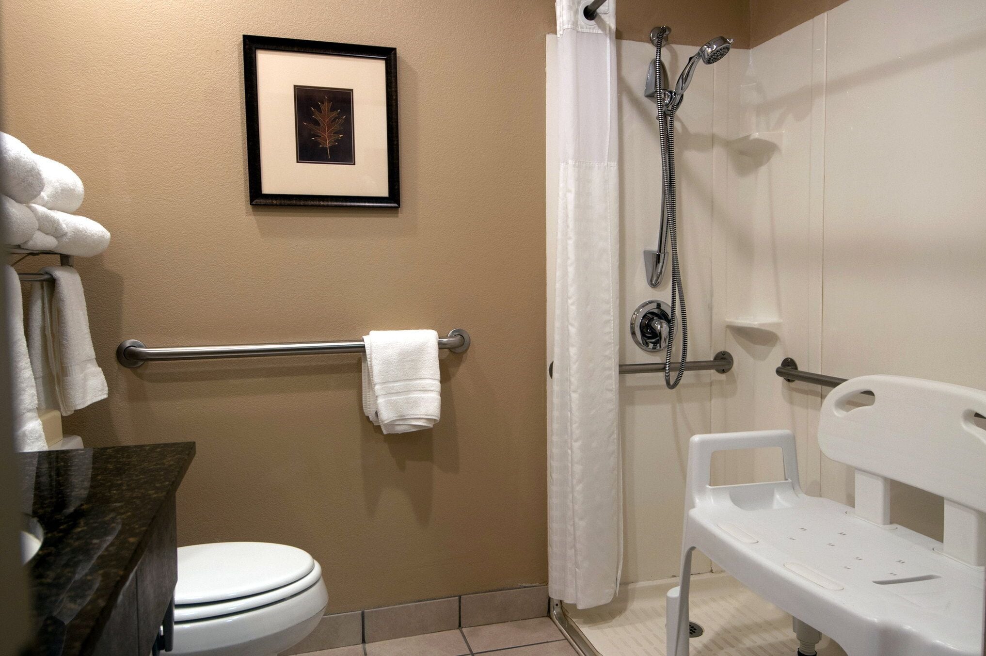 A bathroom with a toilet, a framed picture on the wall, a towel rack with towels, a shower with a handheld showerhead, and a white bench.