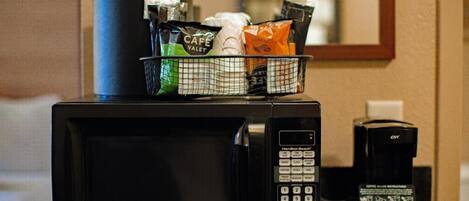 A microwave with a coffee maker and a basket of coffee pods on a countertop.