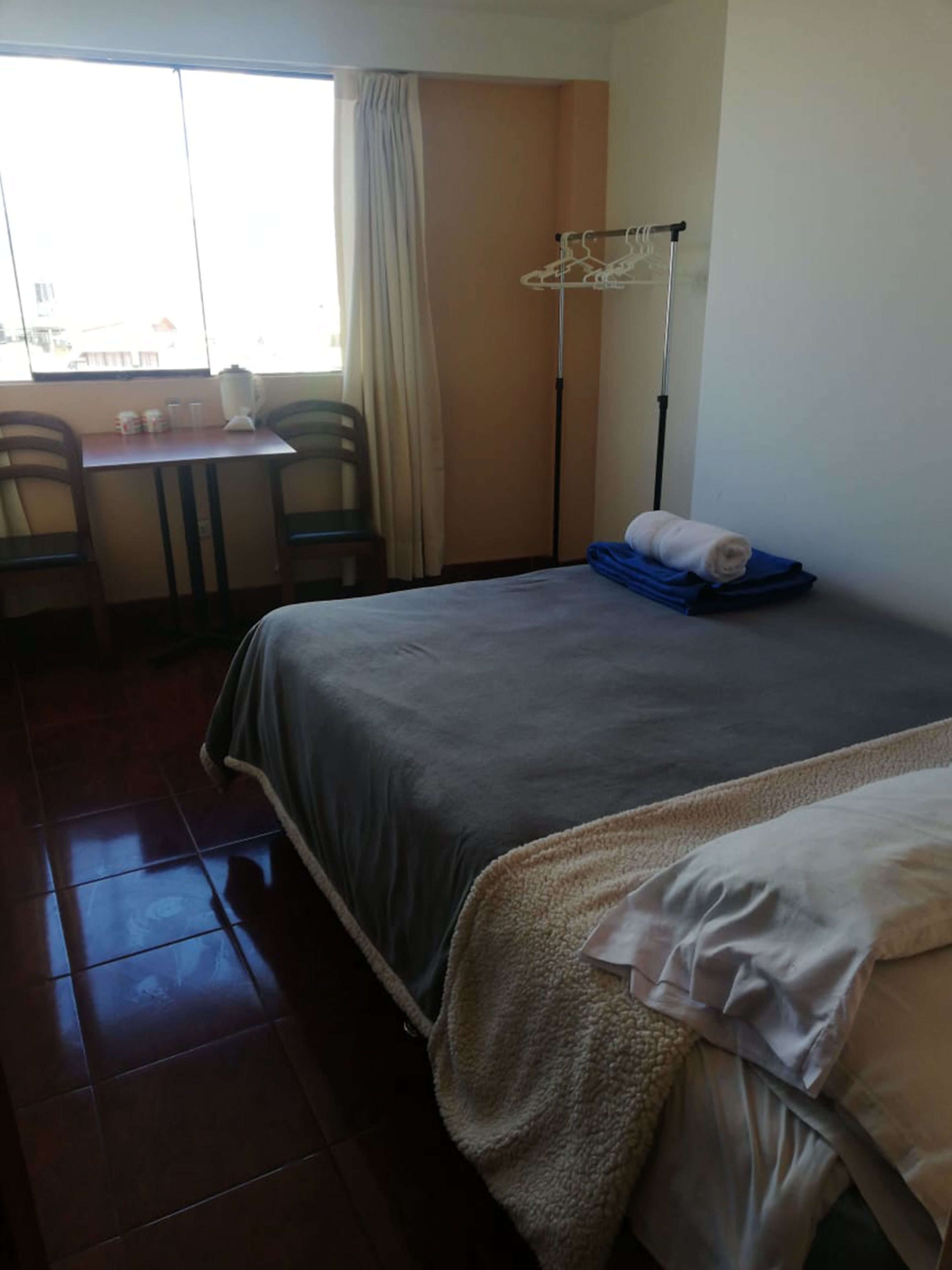 Habitación de hotel con cama, escritorio y silla. Se ve una ventana con cortinas, un radiador y un perchero.