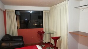 Habitació d'hotel amb un llit, un sofà vermell, dos tamborets vermells, una taula de vidre petita i una finestra amb cortines.