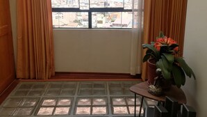 Una habitació amb un gran finestral, cortines, una taula petita amb una planta i un escriptori amb un ordinador i altaveus.