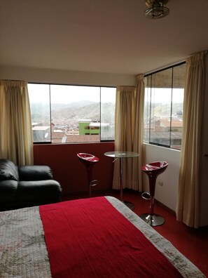 Habitación de hotel con un ventanal grande, una cama, una manta roja, dos taburetes altos y un sillón de cuero negro.