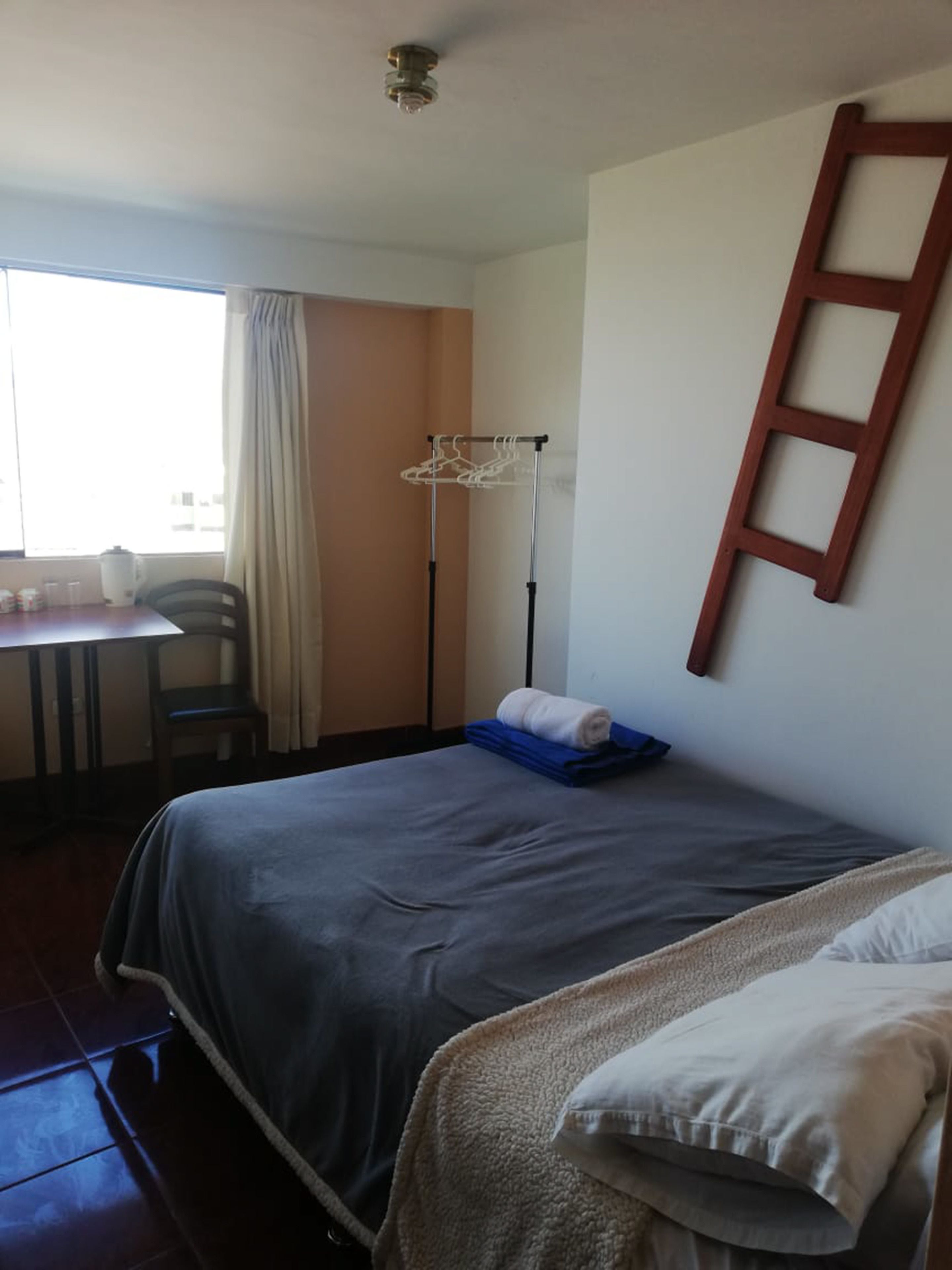 A hotel room with a bed, a desk with a chair, a window with curtains, and a wooden ladder on the wall.