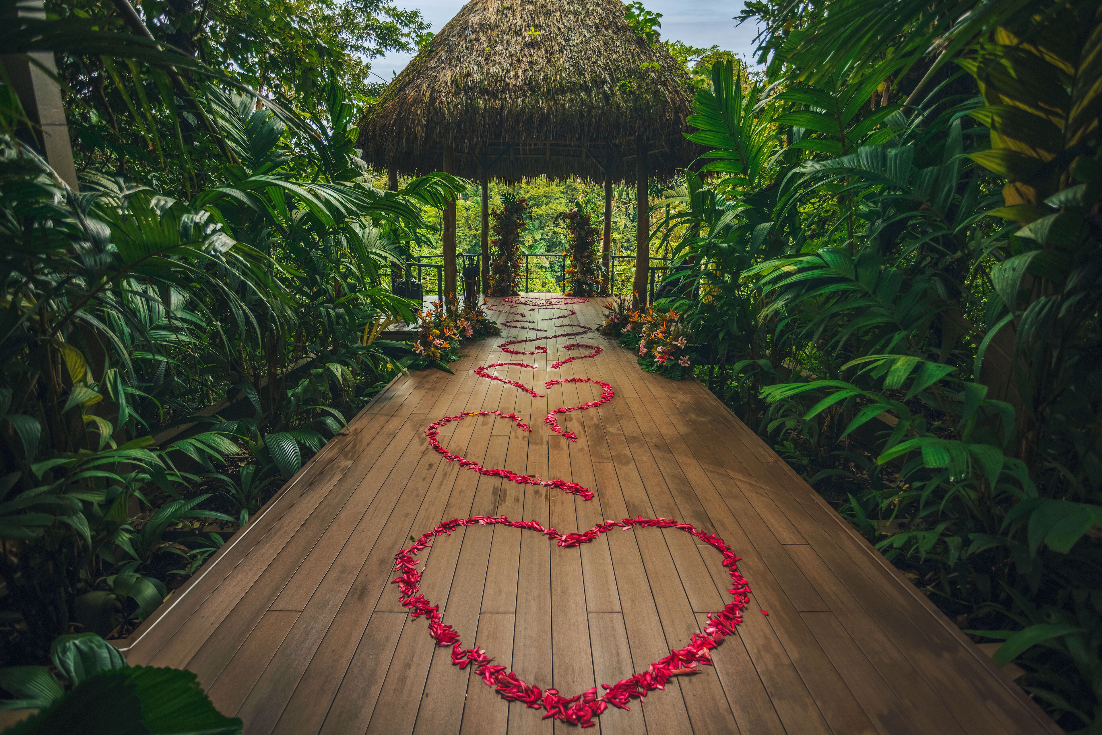A wooden pathway with heart-shaped flower arrangements leading to a thatched-roof structure surrounded by lush greenery.